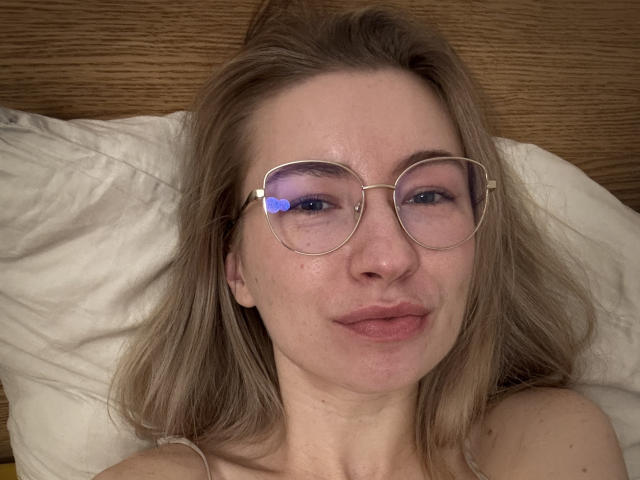 MiaFlirt, con cabello castaño claro, usando gafas con un emoji de corazón azul en la lente derecha, acostada sobre un almohadón blanco, sonriendo ligeramente, mostrando pecas y un maquillaje sutil..