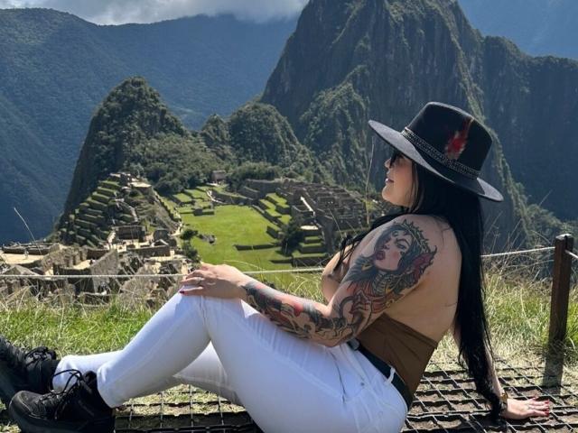 MaeveBorch with tattoos on her arm, sitting on a fence, wearing white pants, brown top, black hat with red feather, black shoes, looking at Machu Picchu.
