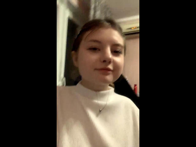 Paris, de pele clara, com cabelo castanho preso em um rabo de cavalo, vestindo uma gola alta branca, nariz pequeno, um leve sorriso e um colar com um pingente minúsculo, em um quarto com pouca luz..