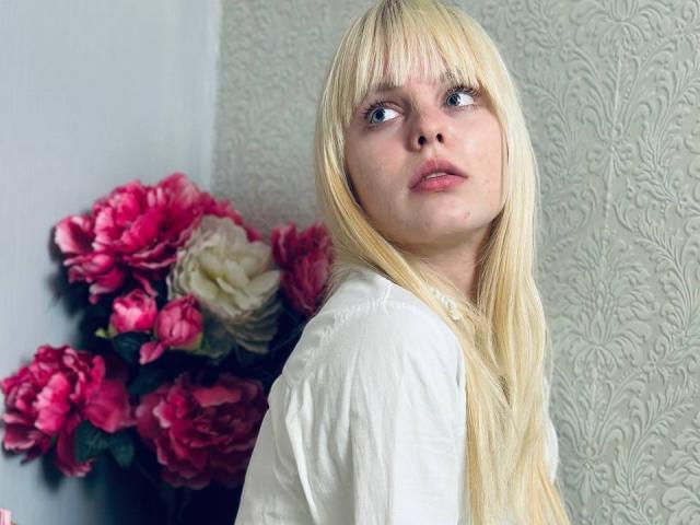 Blonde Lialit met blauwe ogen en een pony, die een witte blouse draagt, kijkt opzij met een licht verwarde uitdrukking, en staat naast roze en witte bloemen..