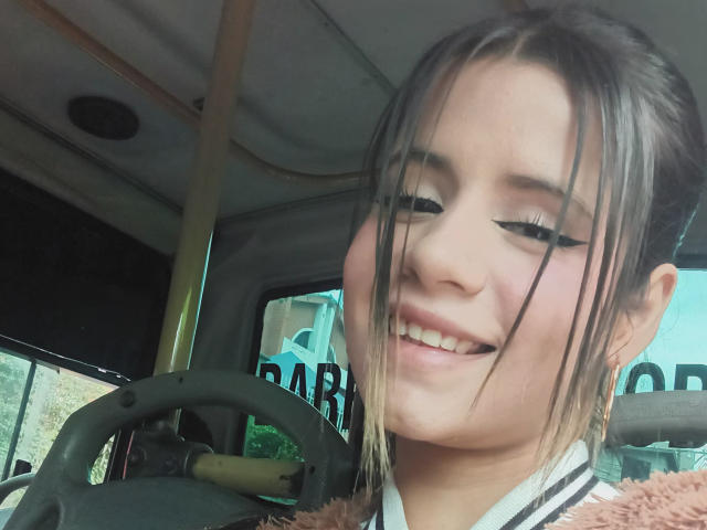 SheylaSpooner smiling with black eyeliner, brown hair in a messy bun, and a fluffy pink jacket, sitting in the driver's seat of a bus.