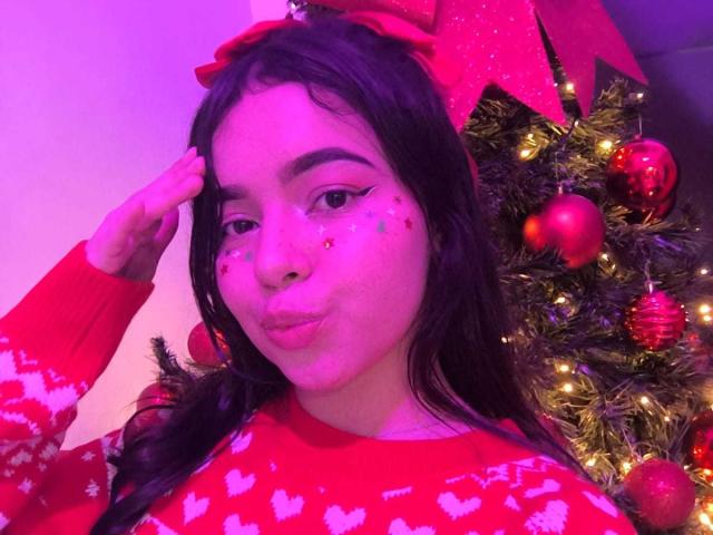 LissyCarter with dark hair, red Christmas sweater, glitter on cheeks, red bow, pouty lips, hand touching hair, Christmas tree with red ornaments and lights in purple-lit background.