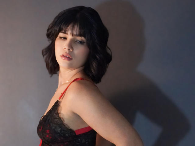ScarlletZhen with short black hair, red and black lace bra, looking seductively to the side, shadow on gray wall, light on left side, slight smile, soft light.