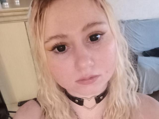 FoxyLunaWs with blonde wavy hair, black eyeliner, and a black leather choker with a heart-shaped metal ring, staring at the camera with a neutral expression.