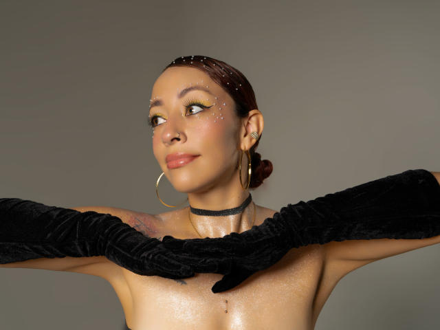 CharlotRey with shiny skin, brown hair, gold hoop earrings, black choker, black velvet gloves, and sparkly cheeks, looking to the side with a subtle smile.