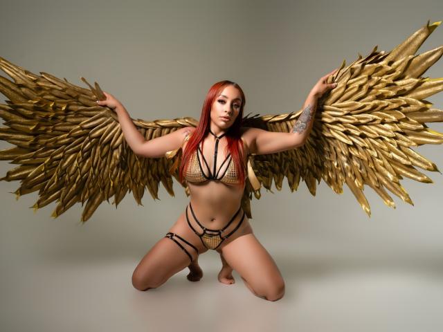 CharlotRey met gouden vleugels, knielend, gekleed in een gouden netbikini en een zwarte string, waarbij haar borsten en vulva zichtbaar zijn, met rood haar en een tatoeage op haar linkerarm..