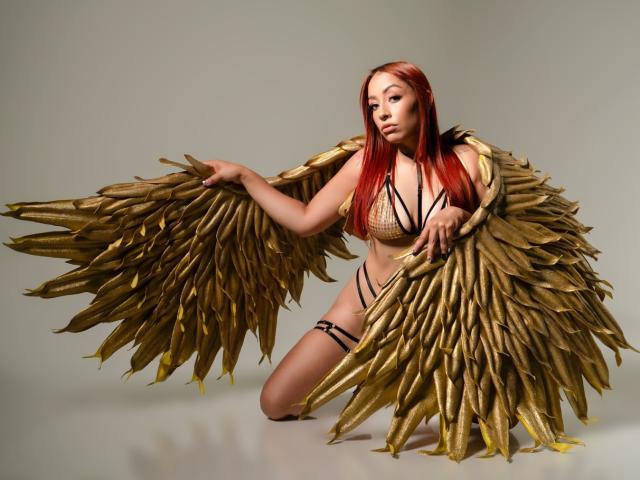 CharlotRey with gold feather wings, red hair, and a gold bralette, kneeling, showing her tits and ass, wearing black strappy panties, holding wings open.