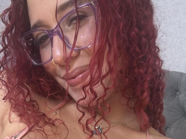 ValeryScot with curly red hair, purple glasses, and a nose ring, smiling slightly, showing cleavage with a necklace, looking into the camera.