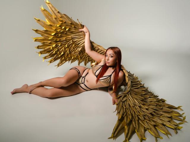 CharlotRey with golden wings, lying down, wearing black mesh lingerie, showing her tits and pussy, with long red hair, on a gray surface, looking at the camera.