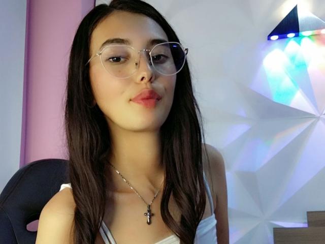 AuraMoon, com cabelos longos e escuros, usando óculos e uma blusa branca, tem um colar com um crucifixo e um leve sorriso, mostrando seus lábios rosados e sua pele clara..