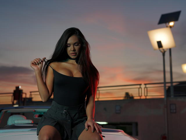 NicolleDiva with long black hair, wearing a tight black tank top and dark shorts, sitting on a car at sunset, holding her hair, showing her tits, with a lamp.