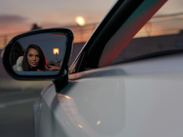 NicolleDiva's face in side mirror, staring out with serious expression, reflected in white car's mirror. She's got long brown hair, and a small nose. Dim streetlight in background.