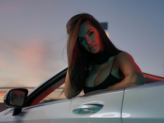 NicolleDiva with long brown hair, red and blue lighting, leaning out of a white car, showing her big tits in a black bra, looking at the camera.