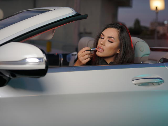 NicolleDiva with brown hair, applying lipstick in a silver convertible with the top down, holding a black lipstick in her right hand, eyes closed, looking sexy.
