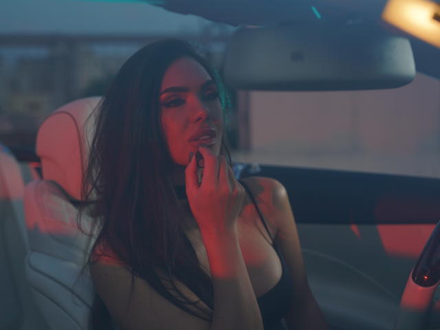 NicolleDiva in a car, red and blue light, dark brown hair, black strap dress, holding cigarette to her lips, white leather seat, serious expression, slightly visible breasts.