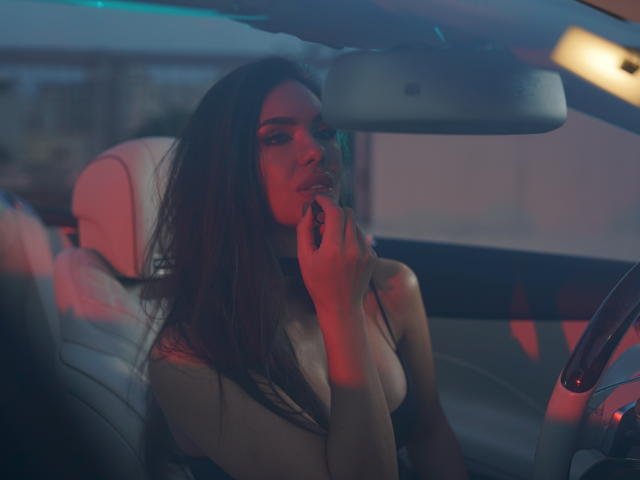 NicolleDiva with long brown hair, sitting in a car, holding a cigarette to her mouth, wearing a beige top, red and blue lights reflecting on her.
