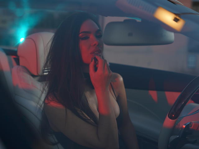 NicolleDiva with long dark hair, wearing a strapless top, holding her phone to her mouth, sitting in a dimly lit car with red and blue lights.