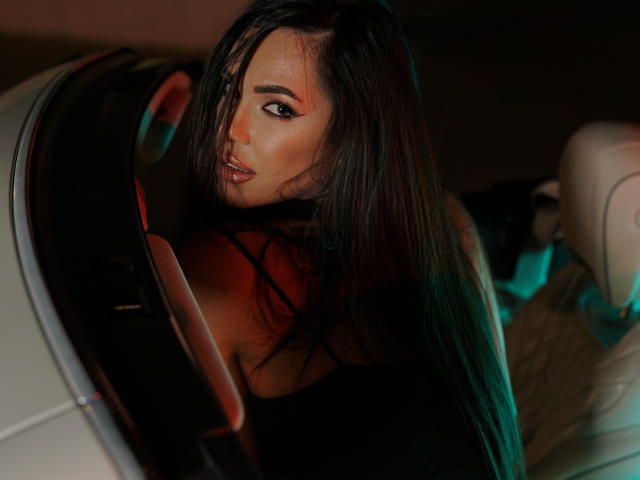 NicolleDiva with long dark hair, looking over her shoulder, in a car, wearing a black top, red light on her cheek, and a sexy, seductive expression.