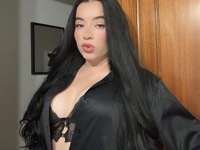 PammelaPrice with long black hair, wearing an open black satin shirt, showing black lace bra with small tits, pouty pink lips, and a neutral expression.