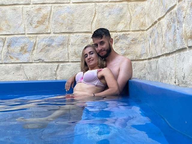 TifanyAndStiven are in a blue pool, Tifany with pink bra, showing her tits, and Stiven shirtless, arms around her, both smiling, water up to their torsos.