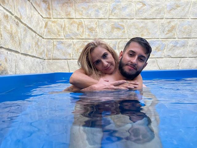 TifanyAndStiven are in a blue-tiled pool, Tifany's boobs are visible above the water, her arms around Stiven's chest, both smiling, with a stone wall in the background.
