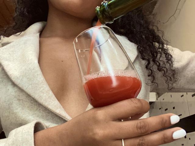 SophieSonyas drinking from a green bottle, pouring red wine into a glass, wearing a gray coat, with curly hair, white nails, and a ring on her right hand.
