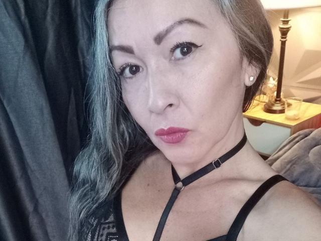 NicoletteShea with gray hair, black choker, and pink lipstick, showing her face and black bra. Her tits are small, and she's wearing a serious expression.