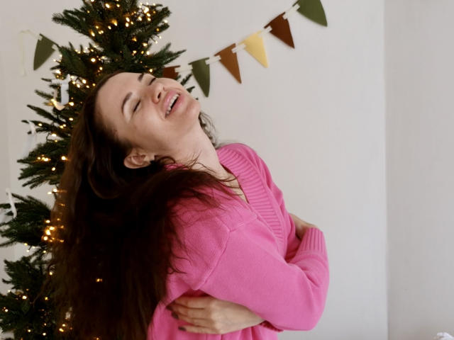 KaraCriegs, com longos cabelos castanhos, olhos fechados, sorrindo, vestindo um suéter rosa, abraçando a si mesma, em frente a uma árvore de Natal com luzes quentes..