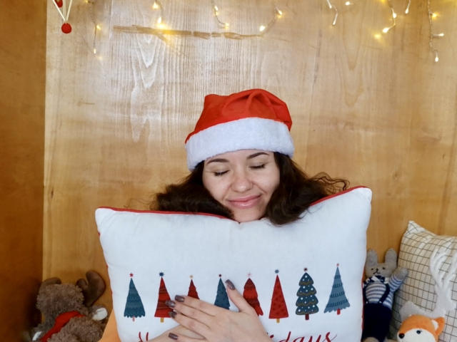 KaraCriegs with curly brown hair, wearing a red Santa hat, smiling while hugging a white pillow with Christmas tree patterns, holding it against her tits.
