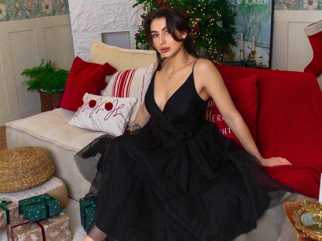OliviaCluff sitting on a red couch, wearing a black, deep V-neck, tulle dress, showing her tits, with wavy dark hair, red lipstick, and a necklace, in a festive room.