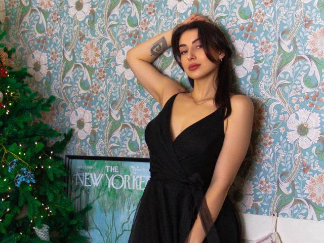 OliviaCluff with dark hair, wearing a black dress, showing her small tits, arm tattoo, hand in hair, standing next to a Christmas tree and a "New York" poster.