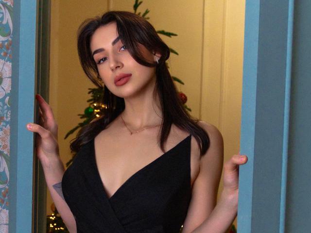 OliviaCluff with dark brown hair, wearing a black deep V dress, holding door frames, showing her tits, with a tattoo on her right hand, and a gold necklace.