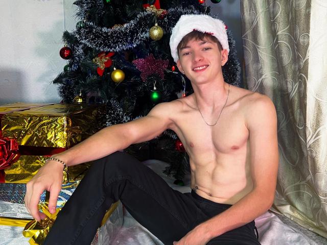 shirtless JakeDavins with a white Santa hat, black pants, and a necklace, sitting in front of a decorated Christmas tree, smiling, showing a toned, muscular, slim body.
