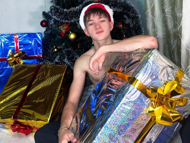 Shirtless JakeDavins with a red Santa hat, holding a silver gift, surrounded by colorful wrapped presents, sitting in front of a decorated Christmas tree.