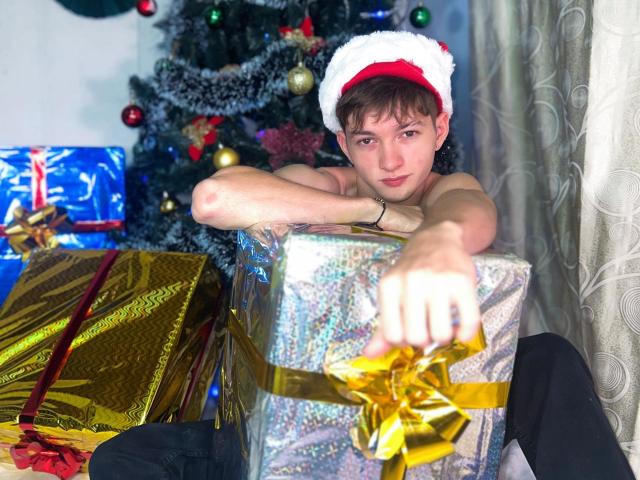 JakeDavins, shirtless, wearing a Santa hat, reaches for a glittery gift with a gold bow, surrounded by colorful presents, looking slightly annoyed. Christmas tree in background.