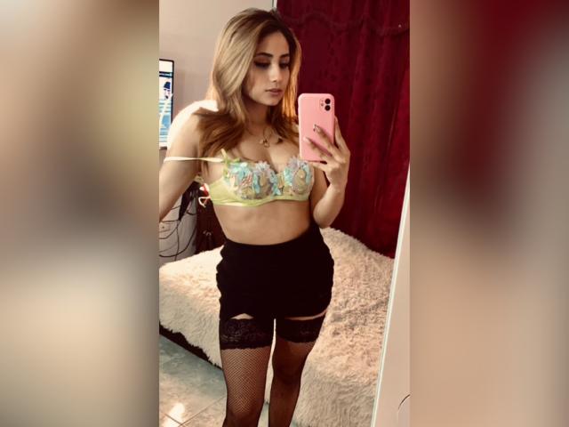 KattyWattson che si scatta un selfie allo specchio, indossando un reggiseno colorato con le tette in evidenza, una gonna nera e collant a rete, tenendo in mano un telefono rosa, in una camera da letto..