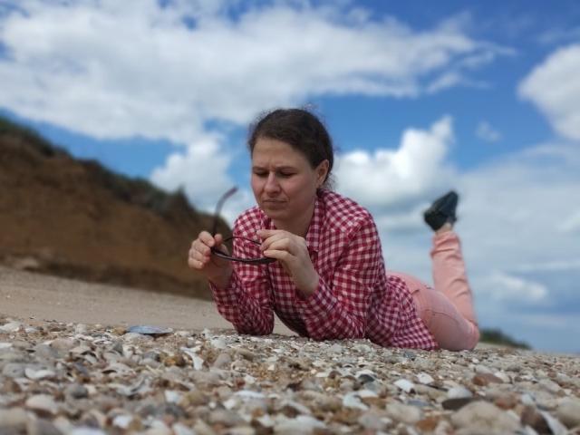 CurlyKsu lying on a rocky beach, wearing a red checkered shirt and pink pants, holding black glasses, with a blue sky and fluffy clouds in the background.