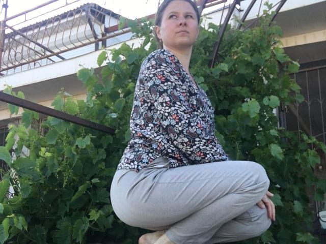 CurlyKsu, squatting with crossed legs, wearing a floral blouse and gray pants, has a medium ass and is looking to the side with a serious expression.