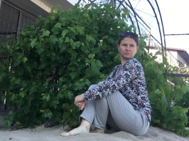 CurlyKsu sits on sand, wearing a floral shirt and gray pants, barefoot, with a green bush behind her and a metal structure overhead. She looks serious.