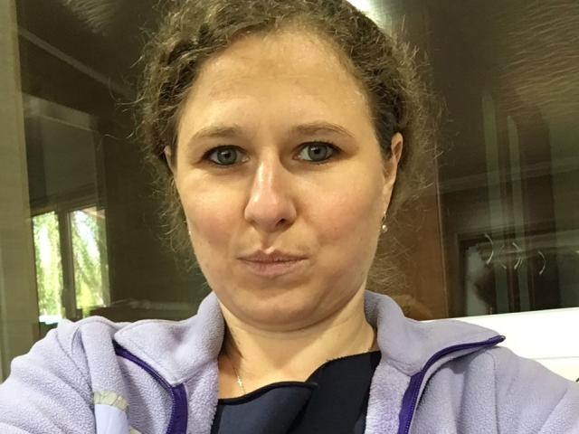 CurlyKsu with curly brown hair, blue eyes, and a pouty face, wearing a purple zip-up hoodie, standing in front of a glass window. Her tits are small.