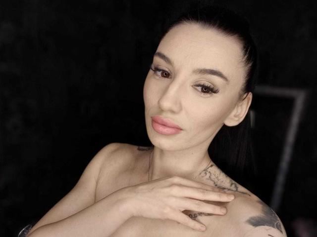 BriannaBratty with pale skin, black hair in a ponytail, brown eyes, pink lips, tattoos on shoulders, arms crossed, looking at camera, dark background.