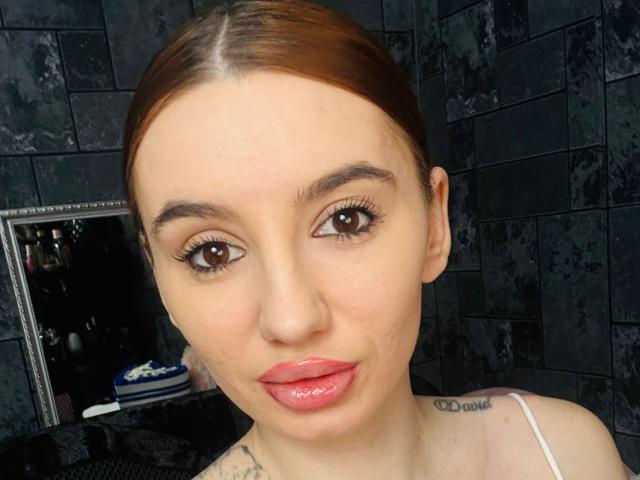 BriannaBratty has shiny pink lips, brown eyes, and brown hair in a slicked-back style, with tattoos on her neck and white spaghetti straps on her shoulders.
