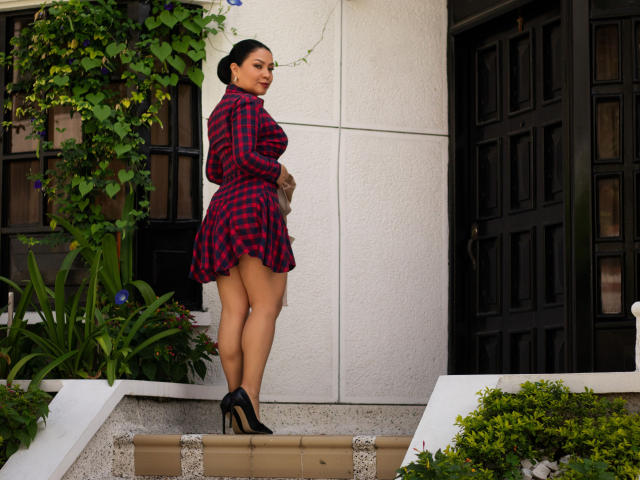 AlisGourmande stands on a porch in black high heels, wearing a red and black plaid dress, showing her ass, with her back to the camera, black door behind.