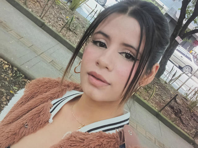 SheylaSpooner with medium tits, brown eyes, and light pink lipstick, wearing a fuzzy pink cardigan and hoop earrings, holding a pink phone, standing outside with trees in background.
