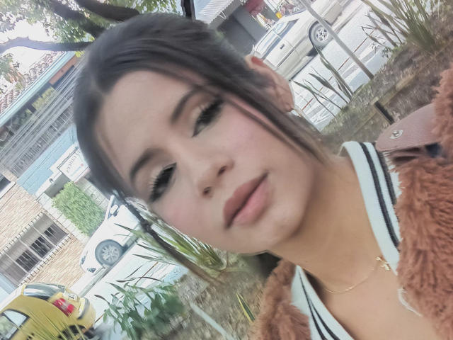 Sheyla Spooner, con labios fruncidos, cejas oscuras y delineador negro, vestida con una chaqueta marrón de pelo, y un collar dorado, de pie al aire libre, con un coche amarillo en el fondo..