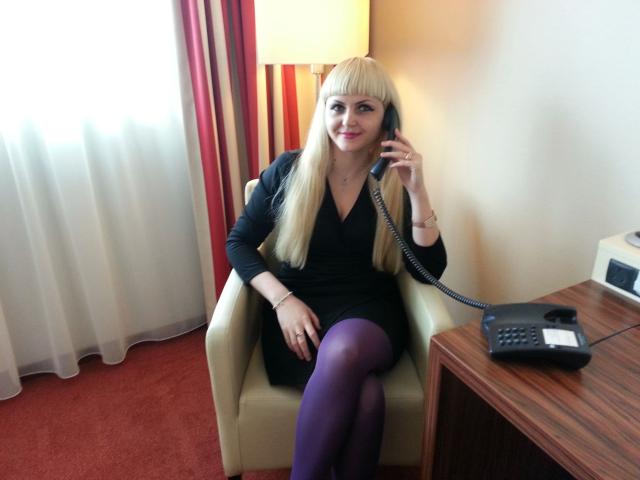 MistresDesiree-hot with blonde bangs, black dress, purple tights, and phone in hand, sitting in chair, smiling, red carpet, white curtain, and wooden table in background.