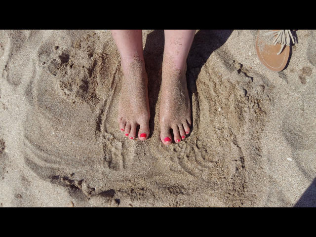 MistresDesiree-hot's sandy feet with bright pink toenails, standing on a beach, and a brown sandal partially visible on the upper right.