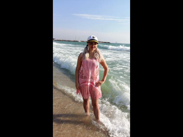 MistresDesiree-hot stands in the shallow ocean, wearing a pink, see-through, fringed dress and a white, yellow-striped cap, with white hair and a confident smile.