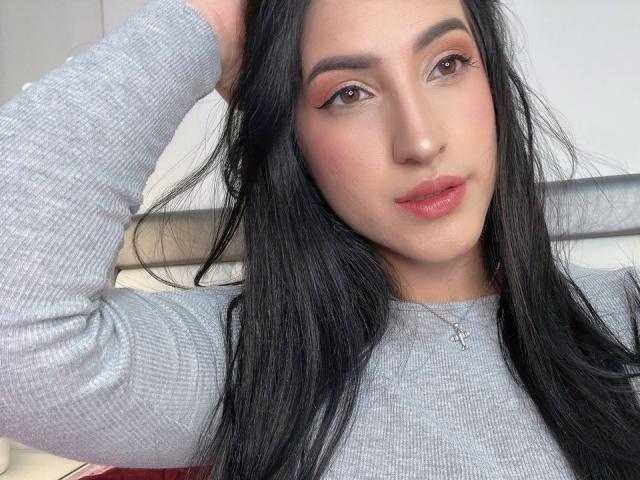 MeganBonnet has long black hair, wearing a gray ribbed sweater, with a small cross necklace, and pinkish-orange eye makeup, her hand in her hair, slightly parted red lips.