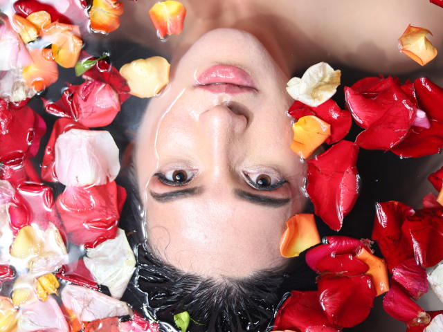 MeganBonnet's face in water with red, white, and yellow petals around her head, looking up with wet hair and a small amount of white fluid on her nose.